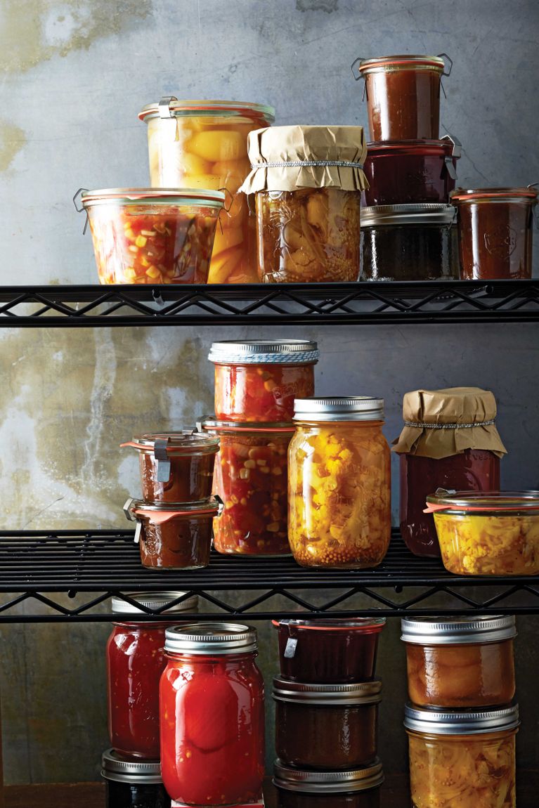 A series of jars of preserves and salsas on black wire racks, including a container of fresh summer salsa made with tomatoes; diced onion; corn kernels; diced red bell peppers; white vinegar; garlic; minced, seeded jalapeño; cilantro; sugar; cumin and salt for a post on an easy fresh summer salsa recipe