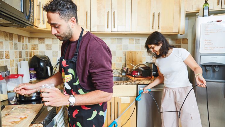 A young couple are in a kitchen. The husband is wearing an apron and cooking at the stove; the wife is vacuuming the floor in the background.