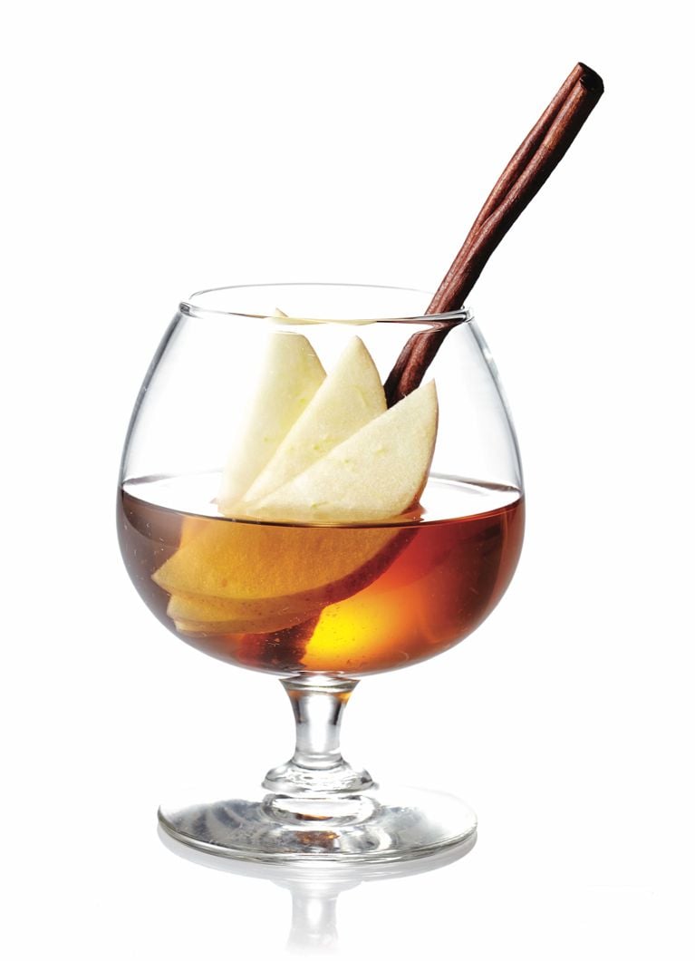 A calvados-vermouth cocktail in a brandy snifter with three fanned slices of apple and a cinnamon stick adorning it for a post on a delicious fall cocktail recipe