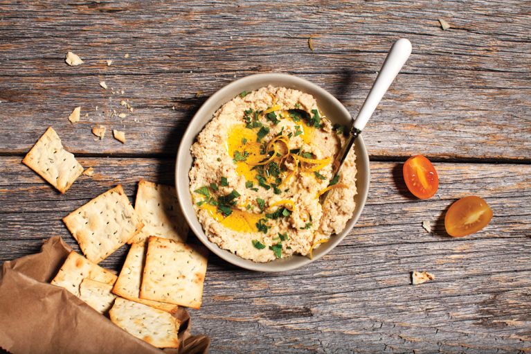 a bowl of cauliflower-tahini dip topped with olive oil and parsley next to crackers and fresh tomatoes for a post on an easy cauliflower-tahini dip recipe