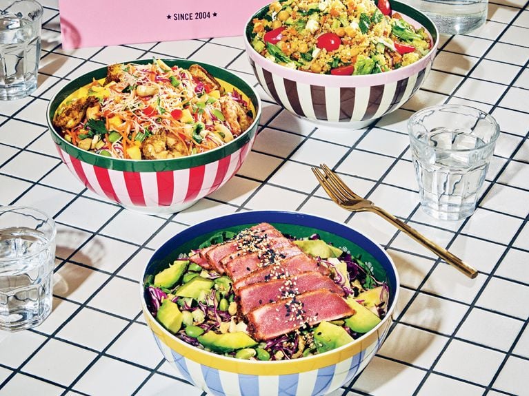 Three bowls of Mandy's salads on a white tile background.