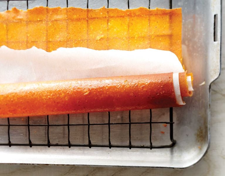 A homemade pumpkin fruit roll up rolled up in parchment paper on a cooling rack; the roll up is made with pumpkin puree; unsweetened apple sauce; pumpkin pie spice and brown sugar for a post on an easy homemade pumpkin fruit rollup snack recipe