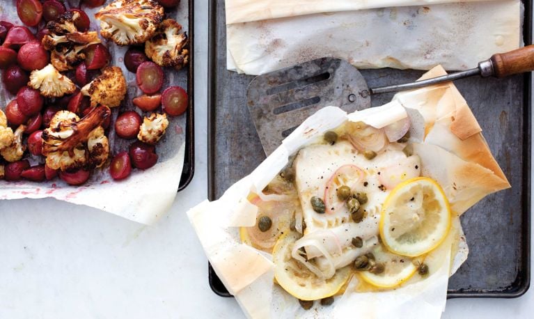 Sheet pan Cod And Caper Parcels With Roasted Cauliflower Salad on parchment paper topped with slices of lemon and a spatula for a post on an easy Cod And Caper Parcels With Roasted Cauliflower Salad recipe for weeknight dinners
