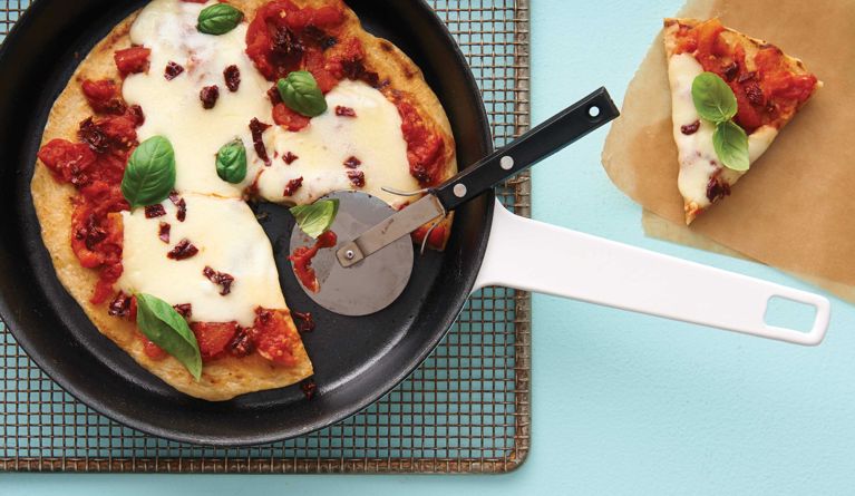 A skillet one-pan pizza in a white pan with a slice cut out of it; the pizza is made with a pizza dough crust; fresh basil; 796-mL can whole tomatoes, drained well; olive oil; garlic; mozzarella; sun-dried tomatoes for a post on an easy fast one-pan skillet pizza recipe