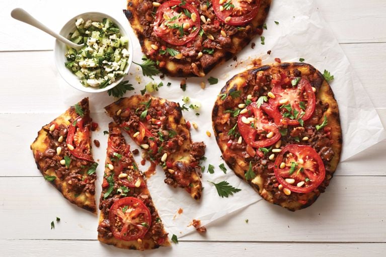 Turkish-style pizza next to a cucumber salad; the pizza is made with naan and topped with ground beef; pomegranate; mint; tomato; pine nuts and parsley for a post on an easy weeknight Turkish-style naan pizza recipe