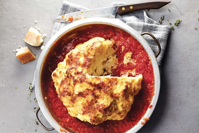 A Whole-roasted cauliflower with tomato-pepper sauce in a pot shown from above next to cubes of parmesan cheese and a knife on its side resting on a kitchen towel for a post on a Whole-roasted cauliflower with tomato-pepper sauce recipe