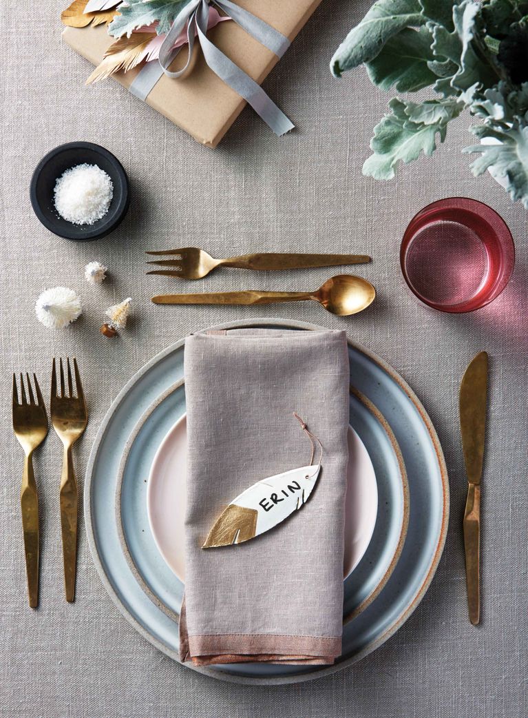 a festive place setting with a napkin with a clay feather ornament dipped in gold with a gold string to hang it and "erin" written on it