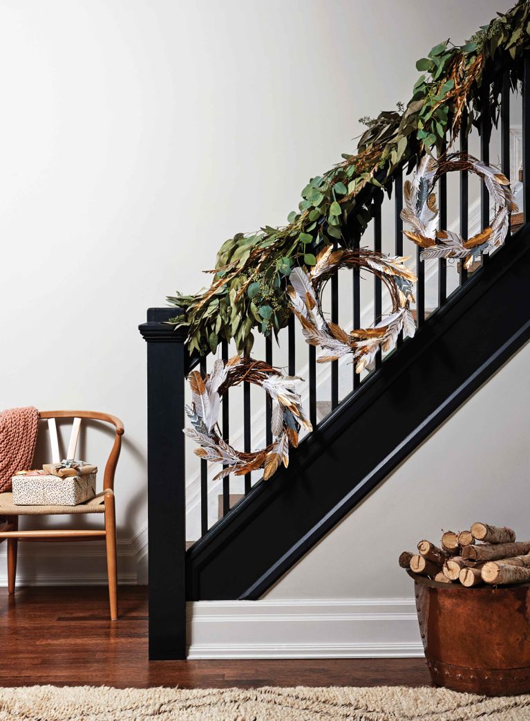 A festive staircase adorned with a green leaves wrapped around the stair railing and white and gold-dipped feather wreaths hanging from the railing