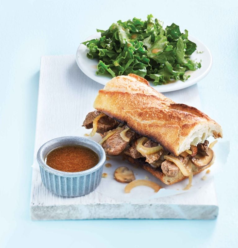 A French beef dip sandwich made with sliced top sirloin steak and mushrooms on a toasted baguette, served with a small side bowl of au jus for dipping and a simple green-leaf lettuce salad.