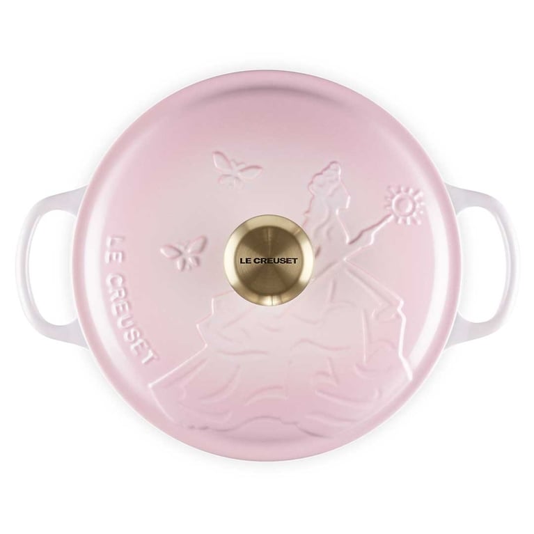 A shell pink Le Creuset dutch oven lid shown from above with an embossing of Glinda from Wicked holding her wand and two butterflies