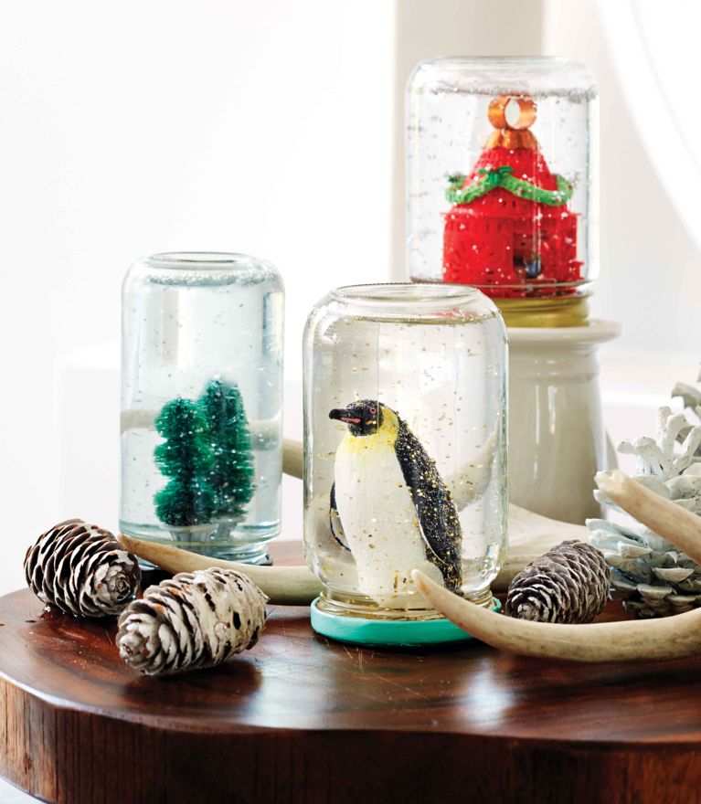 Three homemade snow globes made of figurines or onaments glued to the lid of a glass jar that is then flipped and filled with glitter and water and glycerine for a piece on how to make a homemade snow globe craft or gift
