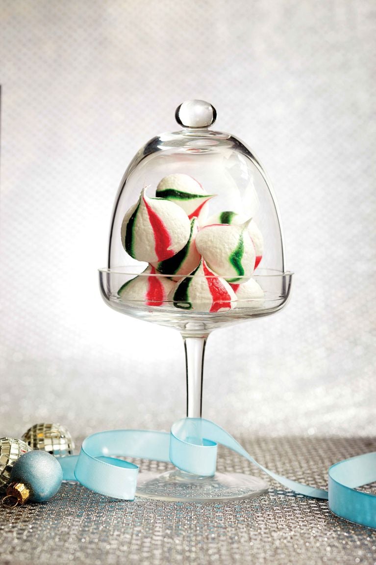 Peppermint Meringue Kisses, made of meringues striped with red and green gel food colouring in a glass serving platter with a glass dome for a post on an easy peppermint meringue kisses recipe