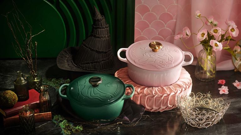 A pink and a green le Creuset dutch oven from the Wicked x Le Creuset collaboration collection. The pink pot rests on a. pink velvet pillow next to Glinda's crown and the green next to Elphaba's pointed hat for a post on the French brand's fall 2025 collections