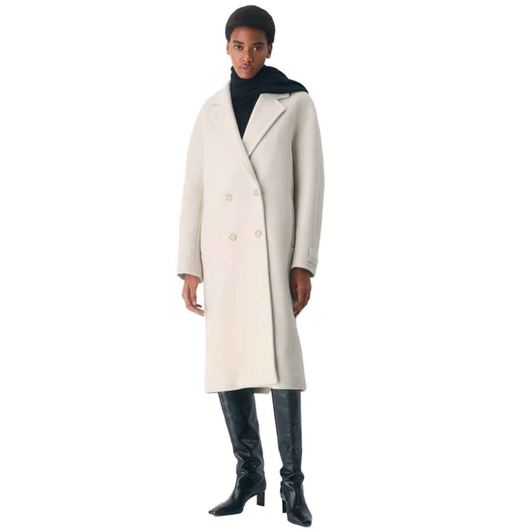 A woman with short hair wearing a long, double-breasted, cream-colored wool coat (the Aritzia Slouch Coat) over a black turtleneck and black knee-high boots with square toes. The woman is standing straight on, looking towards the camera.