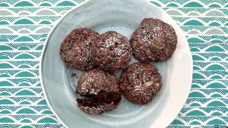 A close-up of several finished Royal's Signature Chocolate Cookies. The dark brown, gluten-free cookies have a chewy, crackly surface and are visibly studded with ingredients like white pumpkin seeds, sunflower seeds, and red dried cranberries.