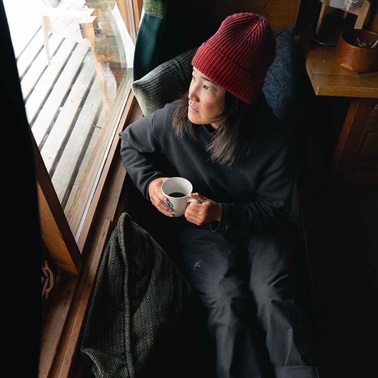 A person wearing a dark grey sweatshirt, dark pants, and a red knit beanie sits on a window seat, looking out a window while holding a white mug with the Arc'teryx logo. There is a dark green textured blanket or sweater draped near them.