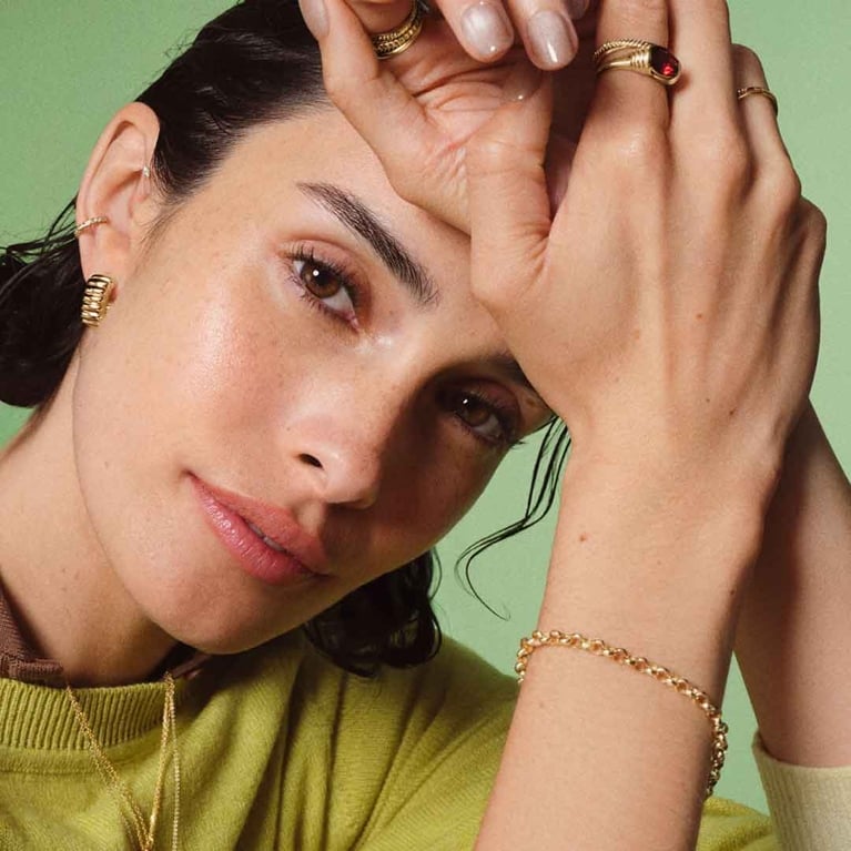 A close-up photograph of a woman wearing a light green sweater and several gold Mejuri jewelry pieces, including thick gold hoop earrings, a delicate chain necklace, stacked rings (one featuring a red gemstone), and a thick chain-link bracelet on her wrist. She is resting her chin on her hands and looking directly at the camera against a soft, green background.