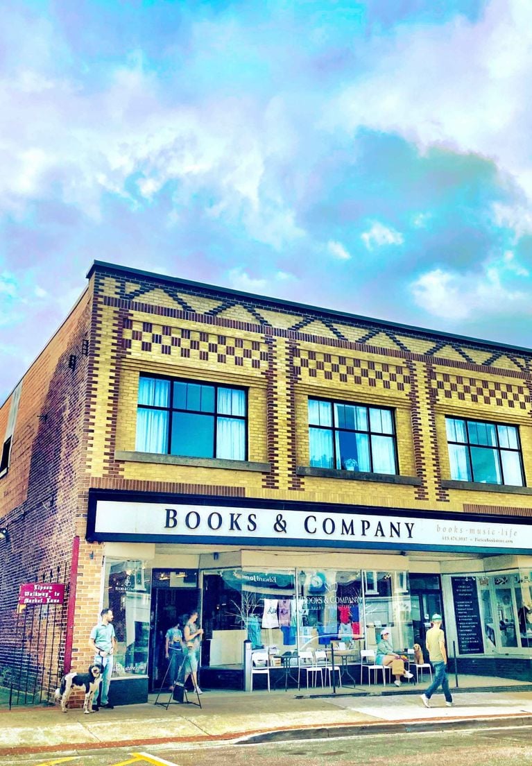 A highly saturated, colorful photo of the exterior of "Books & Company," an independent bookstore in a two-story yellow brick building with ornate trim in downtown Picton, Ontario. The storefront windows reflect the vibrant blue and white sky. People are gathered near the entrance, and one person is walking by on the sidewalk in front.