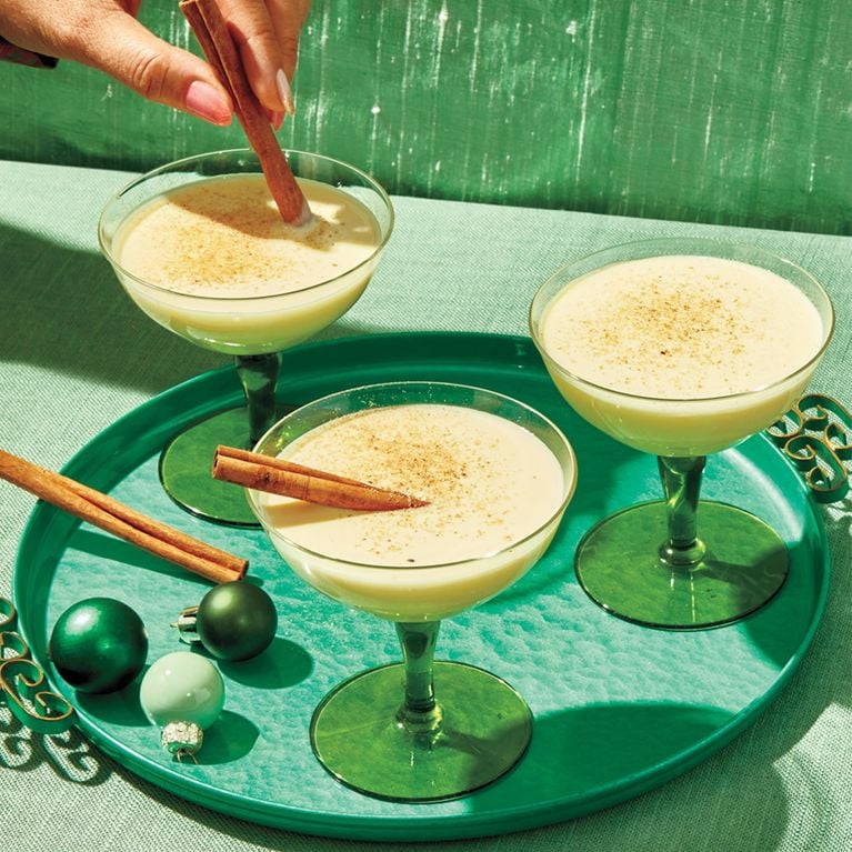 Three coupe glasses of cooked eggnog on a green tray, with a woman's hand stirring a cinnamon stick into one glass.
