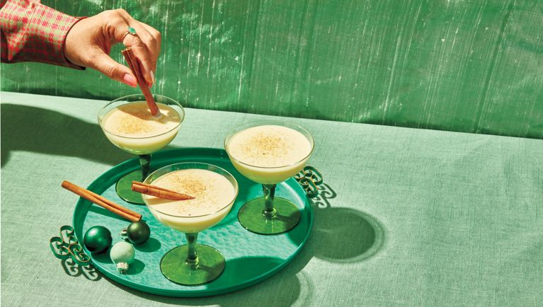 Three coupe glasses of cooked eggnog on a green tray, with a woman's hand stirring a cinnamon stick into one glass.