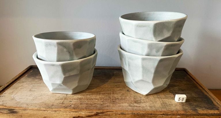 The image shows a set of six handcrafted, faceted ceramic cups with a pale greenish-blue glaze, displayed on a rustic wooden tray or box. Two cups are stacked on the left, and a stack of four cups is on the right. A small white square tag with the number "50" written in black is placed on the wooden surface in the lower right corner.