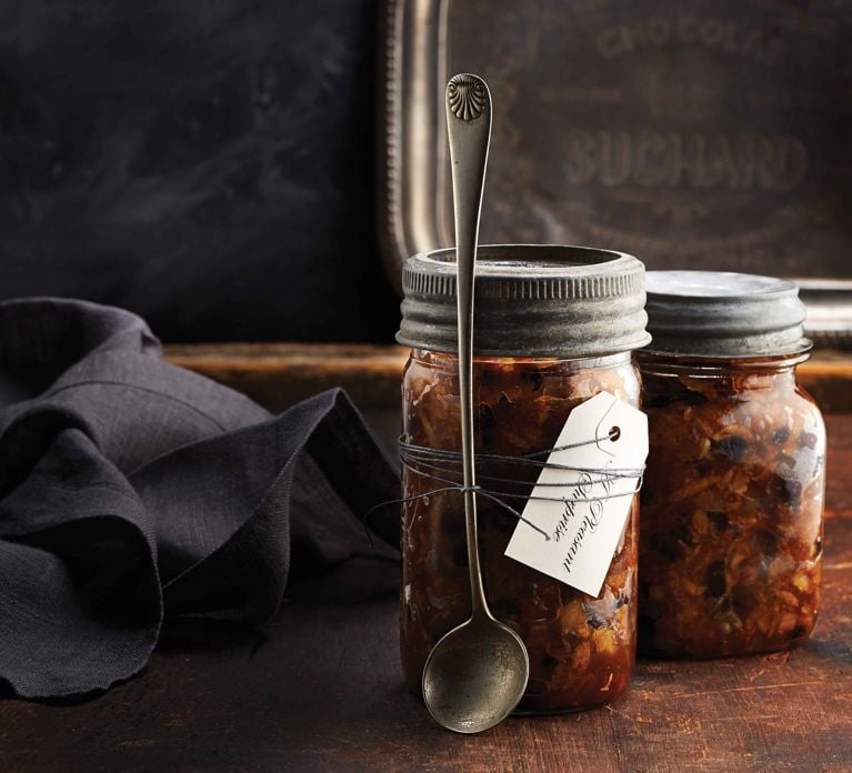 Two vintage glass jars filled with dark, chunky homemade mincemeat, topped with distressed metal lids. A silver spoon rests diagonally across the front jar, which is tied with string and has a white, handwritten gift tag labeled 'Festive Mincemeat'. The jars are set on a dark, rustic wooden surface, with a folded black linen napkin to the left and a tarnished silver tray partially visible in the dark background.