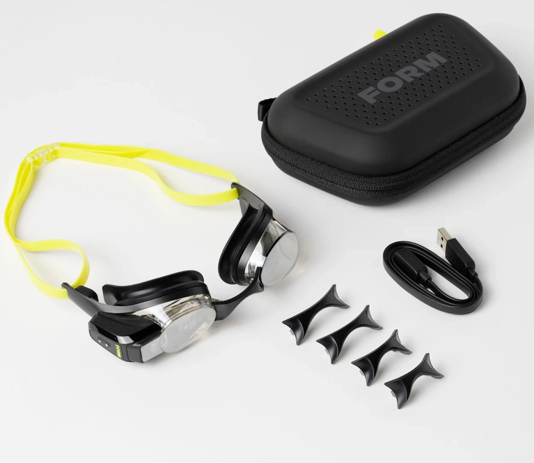 A pair of swim goggles with a yellow band alongside a black case, USB charger and nose fitters, in a post about Form swim goggles.