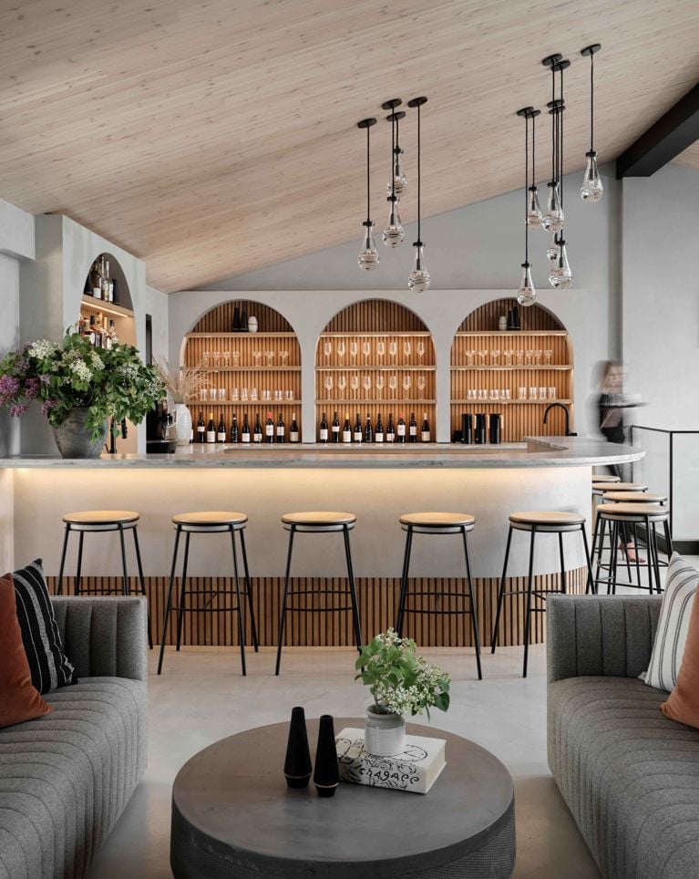 The interior of a stylish, modern bar and lounge area at Gather in Wander in prince edward county. The central bar features a light counter, a slatted wood base, and three arched, backlit alcoves showcasing wine and liquor bottles. The seating area includes a pair of soft grey couches, a round concrete coffee table, and the space is lit by multiple teardrop-shaped pendant lights hanging from a light wood vaulted ceiling.