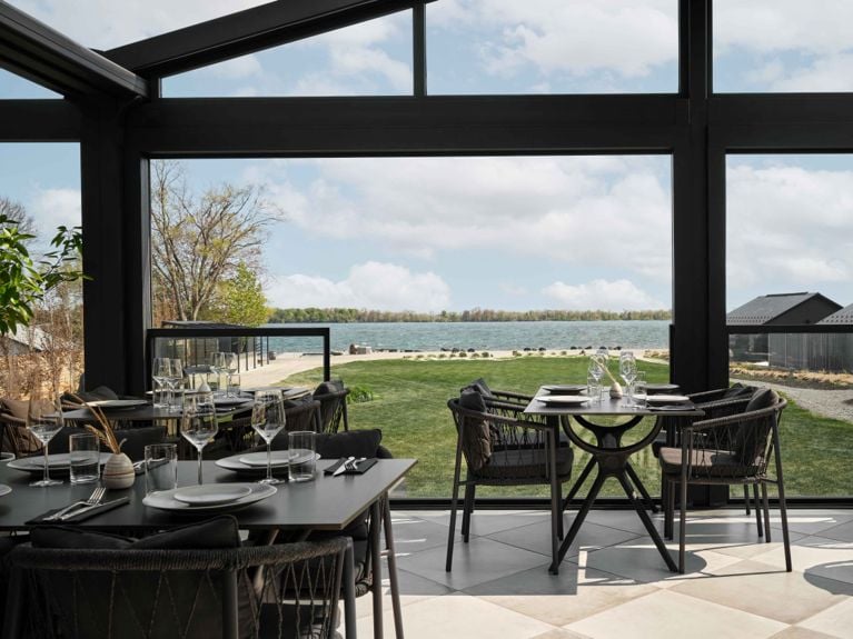 A modern, sunlit restaurant dining area in Gather the spa at Wander with black-framed glass walls looking out over a green lawn and a calm body of water in Prince Edward County. The room features dark, woven dining chairs and tables set with plates and wine glasses, with a light-colored checkered tile floor.