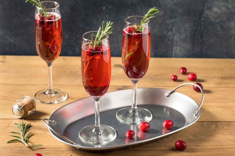 Three champagne flutes filled with a vibrant red cranberry cocktail (Nigella Lawson's Poinsettia cocktail recipe) and garnished with fresh cranberries and a sprig of rosemary, served on a silver tray. A cork and extra cranberries are scattered on the wooden surface nearby.