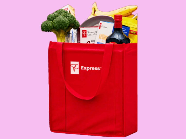 A bright red PC Express reusable shopping bag set against a pink background. The bag is filled to the brim with groceries such as a head of broccoli, yellow bananas, a bottle of dark liquid (wine or sauce), and a box of President's Choice oatmeal. The PC Express logo is visible on the side of the bag.