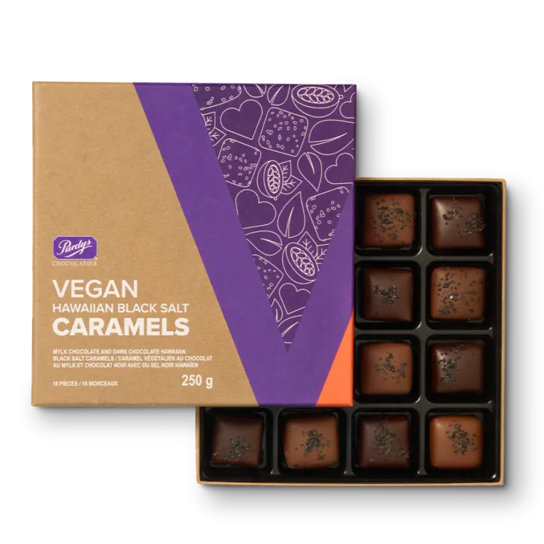 A top-down view of an open box of Purdys Chocolatier Vegan Hawaiian Black Salt Caramels. The box is made of brown cardboard with a purple and orange design on the lid, which features white line art drawings of cacao pods and hearts. Inside the box are 16 square chocolates, dark and mylk, each topped with sprinkles of black salt. The text on the box clearly reads "Purdys Chocolatier," "VEGAN HAWAIIAN BLACK SALT CARAMELS," and "250 g / 16 PIECES."