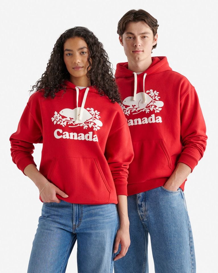 A man and a woman stand next to each other, both wearing identical, bright red Roots "Canada" pullover hoodies featuring the white stylized beaver and logo text across the chest. The woman, with long dark curly hair, is wearing the hoodie with light-wash blue jeans and has one hand in her front pocket. The man, with short dark hair, is wearing his hoodie with medium-wash blue jeans and has both hands in his front pocket. The background is a solid white.