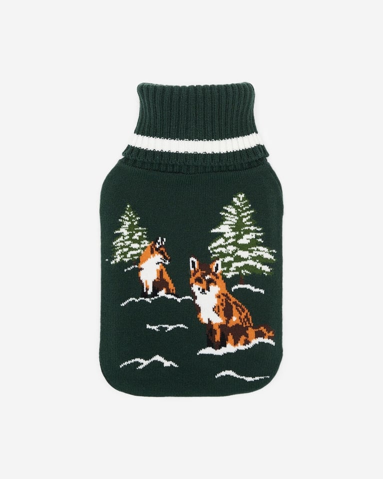 A green knit hot water bottle cover featuring a repeating jacquard pattern. The main motif depicts two stylized orange, brown, and white foxes sitting in a snowy landscape between two white-tipped evergreen pine trees. The neck of the cover is ribbed in a dark green with two horizontal white stripes near the top.