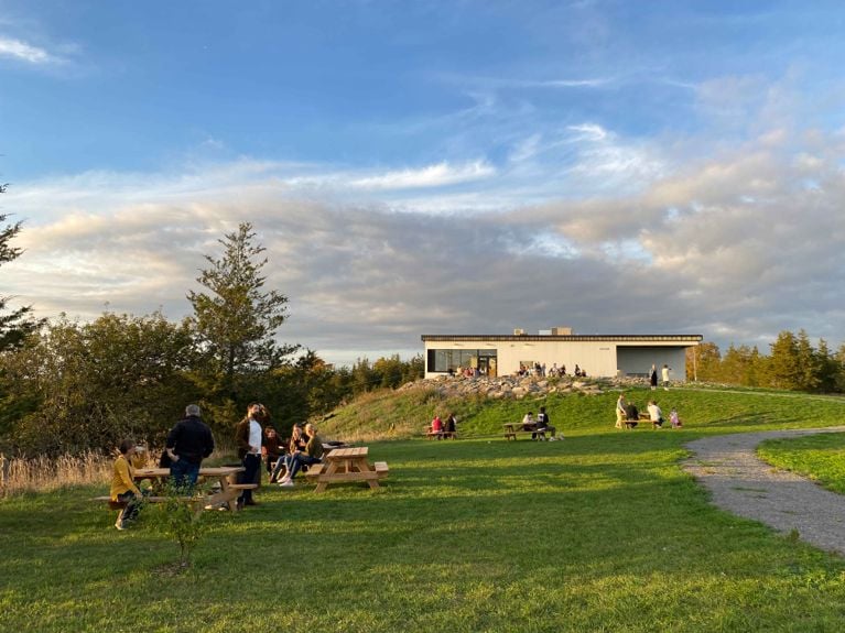 A wide shot of the Slake Brewery building, a modern, minimalist white structure situated on a grassy hilltop. Several groups of people are enjoying the patio and picnic tables on the lawn in the foreground and near the building under a partly cloudy blue sky.