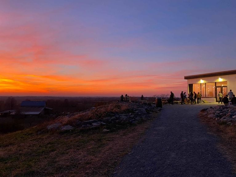 A stunning sunset view from the hilltop at Slake Brewery. The sky is ablaze with vibrant orange, pink, and purple hues. The modern brewery building is visible on the right, and silhouettes of people stand along the gravel path and patio, looking out over the landscape.