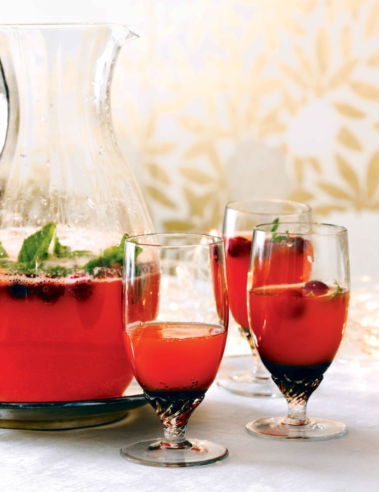 A festive scene featuring a bright red sparkling pomegranate punch. A large glass pitcher filled with the punch, cranberries, and mint sprigs is placed slightly behind three stemmed cocktail glasses filled with the same vibrant drink, garnished with cranberries. The setting is bright with a subtly patterned gold and white background and a white tablecloth, suggesting a holiday party or celebration.