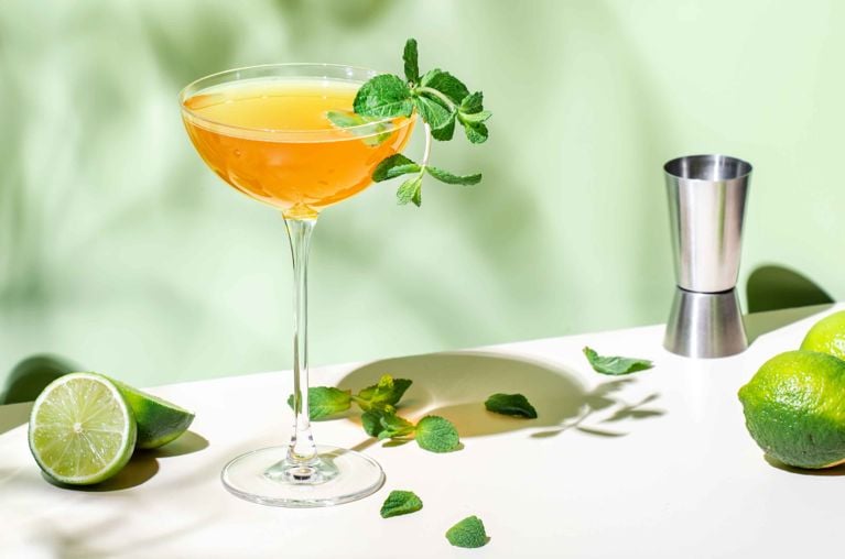 A vibrant, light orange sparkling whisky cocktail is served in a tall-stemmed coupe glass, garnished with a sprig of fresh mint. The glass sits on a white surface next to a jigger and several whole and halved limes, with loose mint leaves scattered around. The background is a soft, pale green, and the scene is lit by bright sunlight casting crisp shadows, creating a fresh, summery atmosphere.