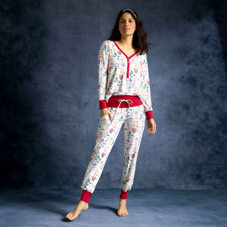 A woman with long, dark hair is standing barefoot and posing against a dark gray/blue studio background. She is wearing a two-piece pajama set with a colorful holiday print (white background with small illustrations of winter scenes and items). The pajamas feature a long-sleeve henley top and jogger-style bottoms, both with red trim at the cuffs and collar/waistband. The woman has her hands in the pockets of the pants, and she is also wearing a matching headband. The lighting highlights her and the texture of the backdrop.