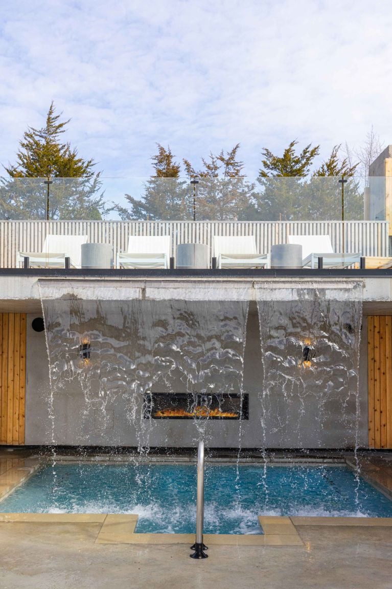 A tall, wide waterfall cascades over an outdoor fireplace and into a spa plunge pool below. The pool has a stainless steel railing for access. Above the waterfall, a row of white loungers sits on a sun deck with a glass railing, backed by low evergreen trees under a bright blue sky.