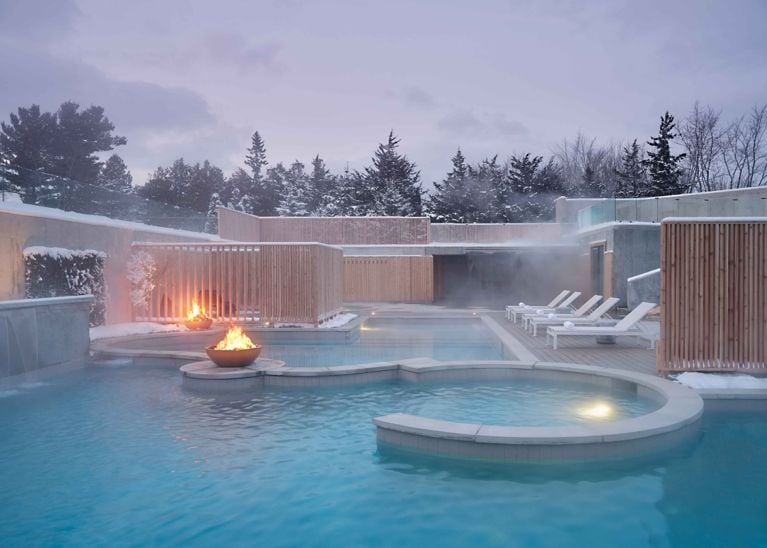 A wide view of the steam rising from the multi-level outdoor spa pools at Wander Resort on a cold, overcast day. The pools are surrounded by light-colored wood privacy screens and a spacious wooden deck with several white loungers. A fire bowl sits on the deck between the winding pools.