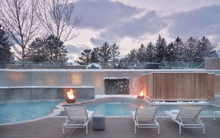 An outdoor Nordic spa pool area at dusk at Wander the Spa in Prince Edward County on a snowy winter day. The multiple heated pools are surrounded by wooden decking and snow-covered evergreen trees. Two fire bowls on raised pedestals flank the pools, casting a warm glow and adding contrast to the cool blue pool water and snowy landscape.