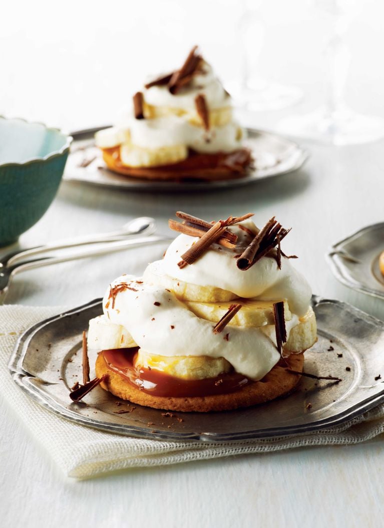 A vertical, high-angle food photography shot of an elegant individual banoffee tart served on a vintage silver plate. The dessert features a crisp, round, golden-brown biscuit base topped with a layer of smooth caramel. Slices of fresh banana are stacked beneath a generous, billowy mound of white whipped cream. The cream is garnished with dark chocolate shavings and curls. In the softly blurred background, a second identical tart sits on a matching plate, alongside silver dessert spoons and a partial view of a blue bowl.