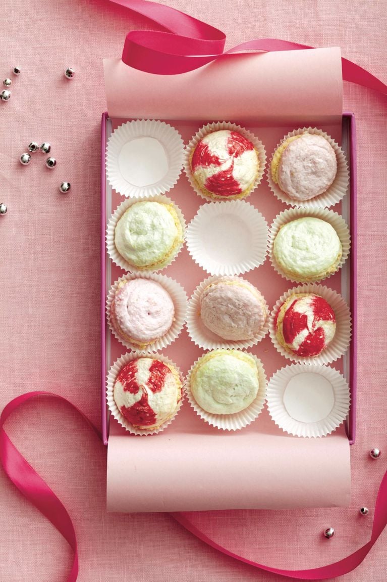 An overhead view of an open pink gift box containing colorful meringue sandwich cookies nestled in white fluted paper liners. The box holds a mix of pale green, soft pink, and white-and-red swirled meringues. Three of the twelve liners are empty, showing only the white paper cups. The box rests on a textured pink fabric surface, decorated with a curling dark pink satin ribbon and scattered silver sugar beads.