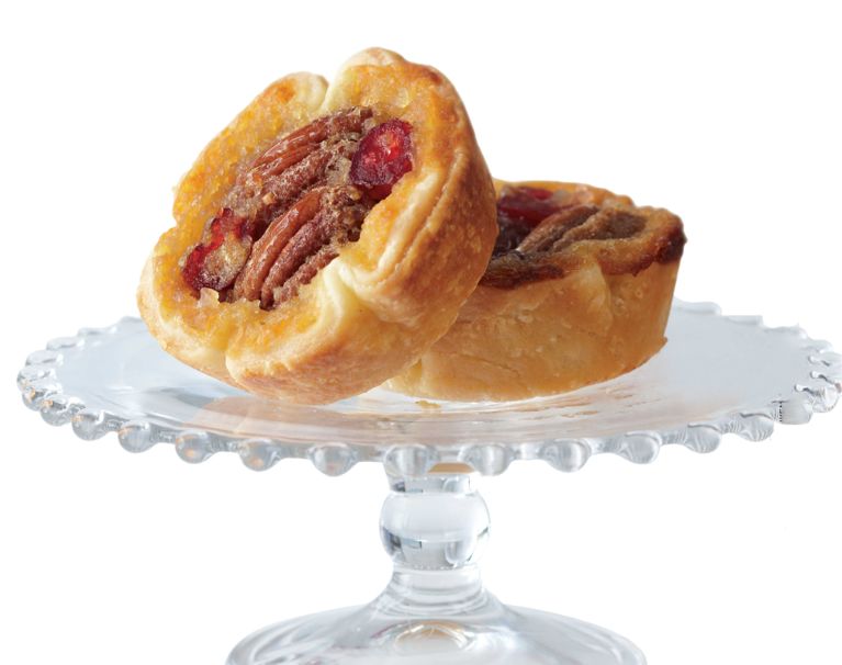 Two golden-brown butter tarts filled with pecans and cranberries resting on a clear glass cake stand with a beaded rim. The front tart is tilted on its side against the second tart, showcasing the flaky pastry crust and the glossy, nut-and-fruit filling against a stark white background.