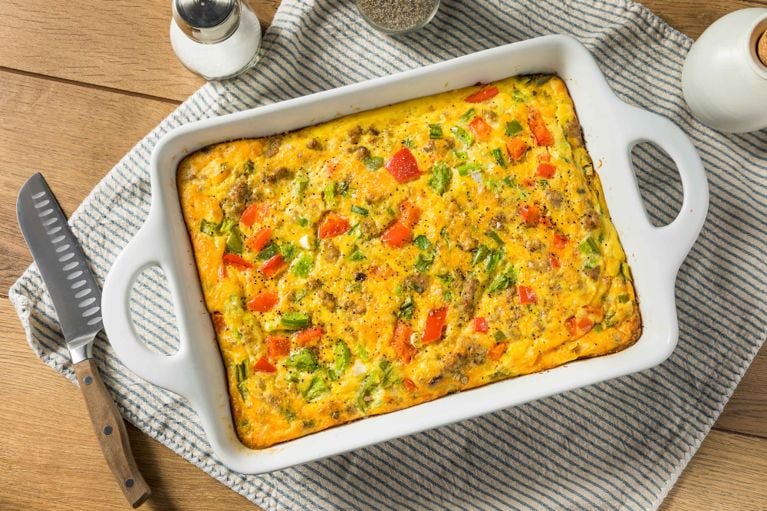 A top-down shot of a golden-yellow egg and cheese bake (also known as a breakfast casserole or frittata) in a white rectangular baking dish. The baked dish is speckled with visible ingredients, including browned sausage crumbles, bright red diced bell peppers, and green diced bell peppers. It has a slightly irregular, bubbly top texture. The baking dish is placed on a light blue and white striped cloth napkin on a light brown wooden surface. To the left of the dish, there is a serrated utility knife with a wooden handle. In the upper left corner, there is a small white salt shaker and a glimpse of a clear bowl containing black pepper. In the upper right corner, there is a white ceramic container (possibly a pepper cellar or a small cream pitcher).