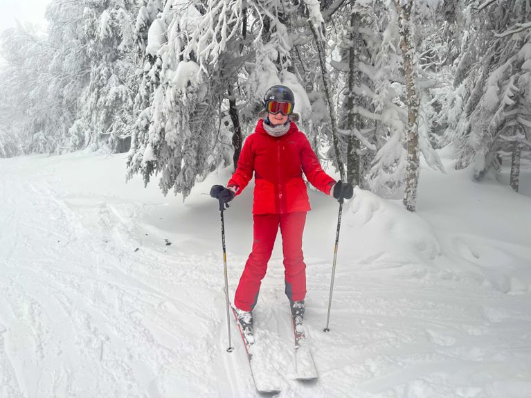 A medium shot of a smiling skier in a red outfit standing stationary on skis in a snowy forest.