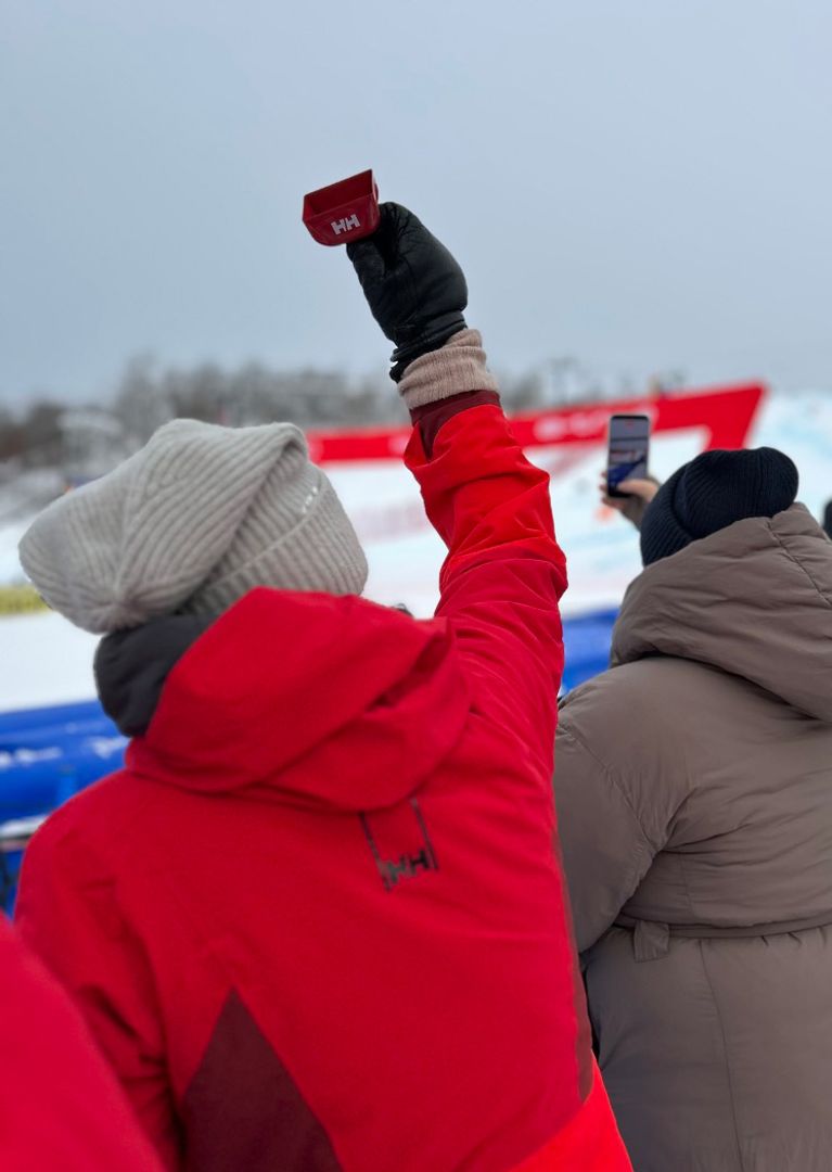 A spectator in a red jacket raises a red cowbell in the air while watching a ski event.