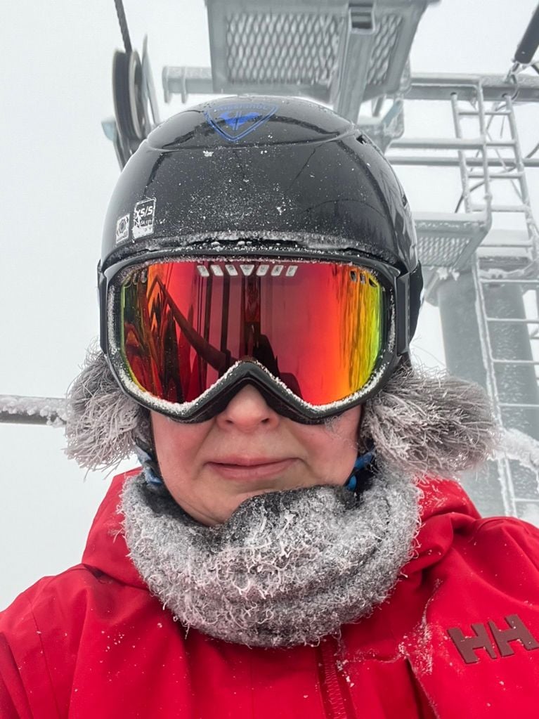 A close-up selfie of a skier on a chairlift, wearing a black helmet and red jacket. Their grey neck warmer and furry ear flaps are covered in frost and ice crystals.