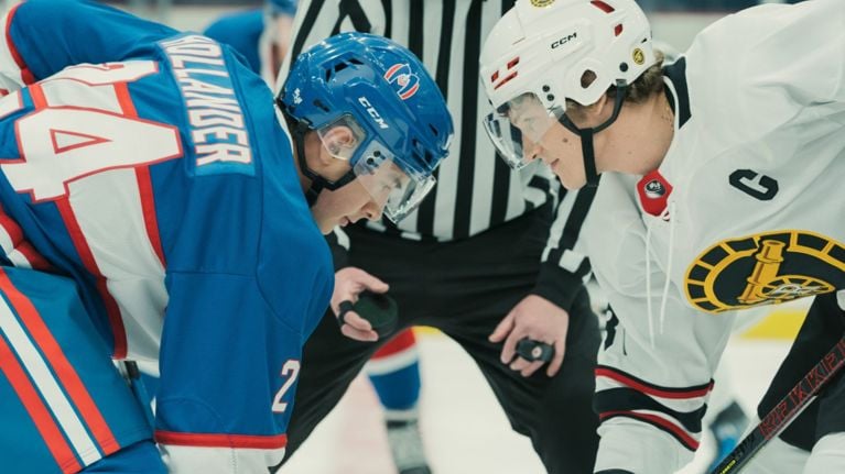 Two young hockey players (Hudson Williams as Shane Hollander and Connor Storrie as Ilya Rozanov) prepare for a face-off with a referee and other players visible in the background.